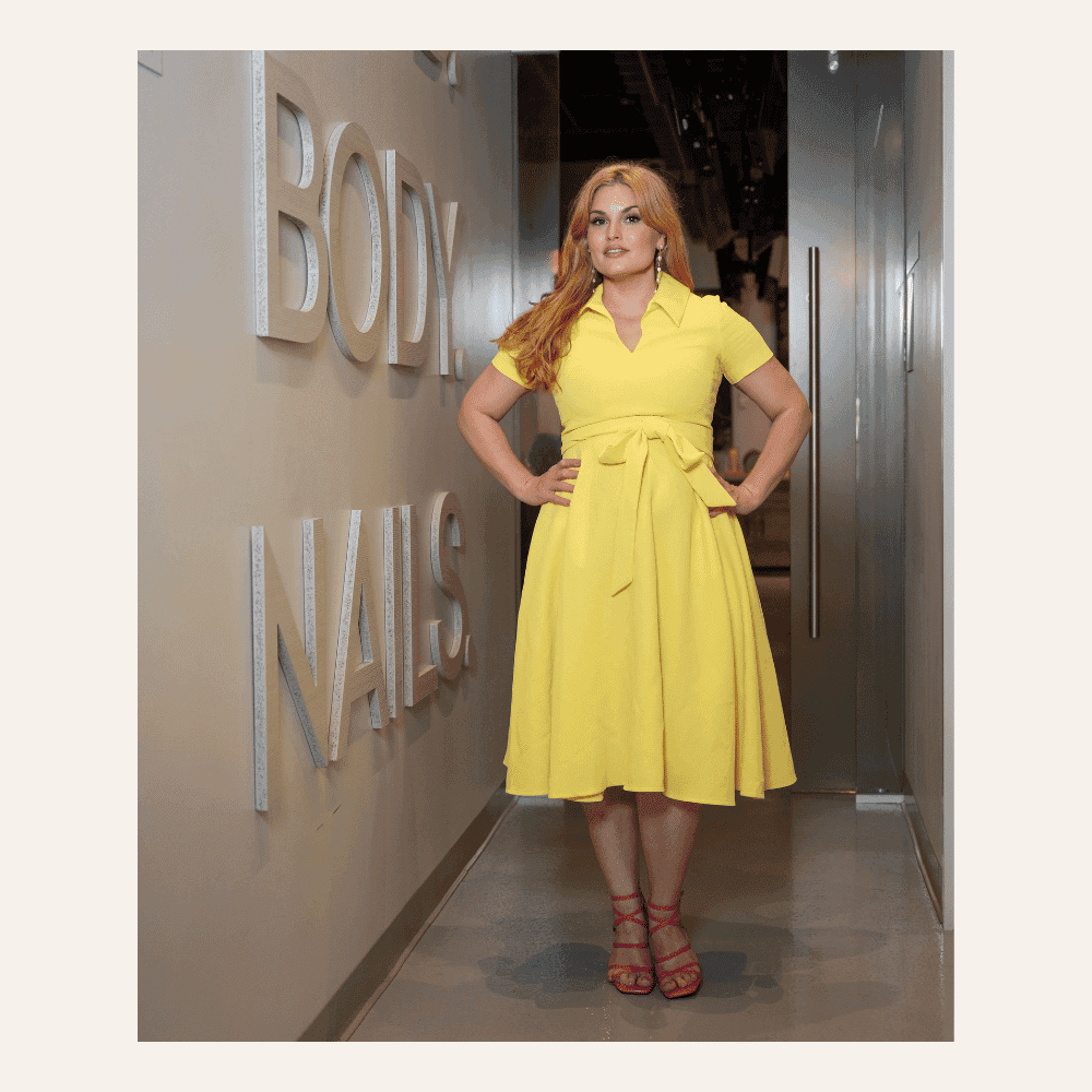 Model in yellow dress with pink heels stands confidently in hallway near wall signage.